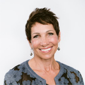 A woman with short, dark hair smiles brightly, wearing a grey flowered sweater.
