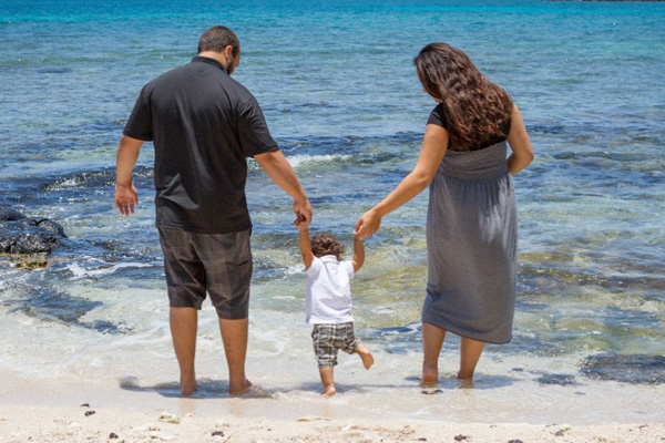 A man and a woman each holding the hand of a toddler while they walk through clear beach water.